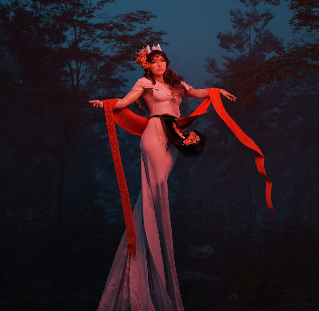 Paulina del Campo in a flowing mexican dress stands amidst a misty forest holding a red ribbon and a floral accessory with a serene expression on her face.
